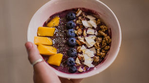 Hand holding a bowl of acai smoothie topped with mango slices, blueberries, chia seeds, coconut flakes, and granola.