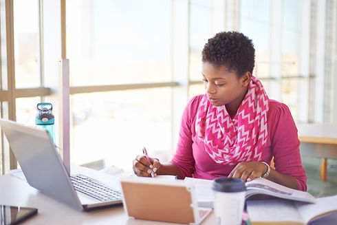 A person in a pink shirt and chevron scarf studies at a table with a laptop, notebook, and coffee cup