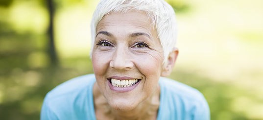 Smiling older adult with short white hair in a light blue shirt, leaning toward the camera outdoors with a blurred green background.