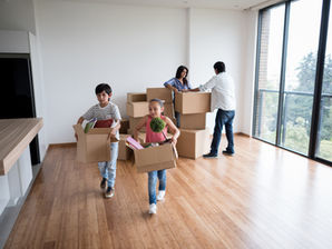 Family moving into new home, two kids carrying boxes in the foreground, parents organizing in the background, bright room, wooden floor.