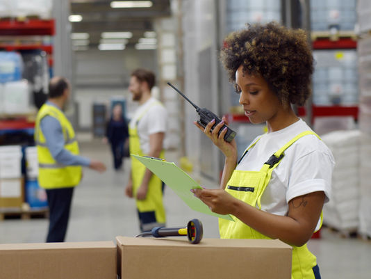 Warehouse worker in neon vest checks a clipboard and handheld radio among stacked boxes, with coworkers blurred in the background.