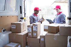 Two men in red shirts and hats stand beside stacked boxes, engaged in conversation