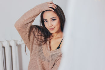 Smiling woman with long brown hair in a cozy beige sweater poses playfully, with a bright, blurred background emphasizing a warm, relaxed mood.