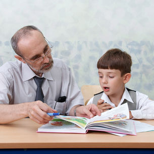 A man helps a boy read a colorful book at a desk. The setting is a classroom with a light abstract wall pattern, creating a learning atmosphere.