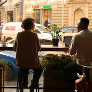 Two people sit in a cafe by a window, with plants and a laptop on the table. Outside, cars pass. Warm lighting, cozy atmosphere.