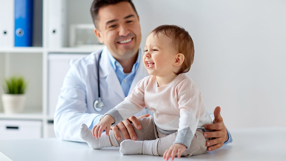 Smiling baby in pink and gray sitting on a white table, held by a doctor.