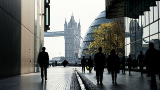 Silhouetted people walk between modern buildings in London. Tower Bridge and City Hall are visible. Reflections and soft light create contrast.