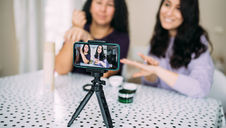 Two women recording a video with a smartphone on a tripod on a polka dot tablecloth. Both smile as they demonstrate skincare products.