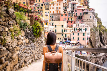 A traveler with a leather backpack takes in the vibrant, colorful hillside buildings of Manarola, Italy, on a sunny day.