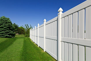 A long, white picket fence stretches diagonally through a lush, green lawn with a clear blue sky