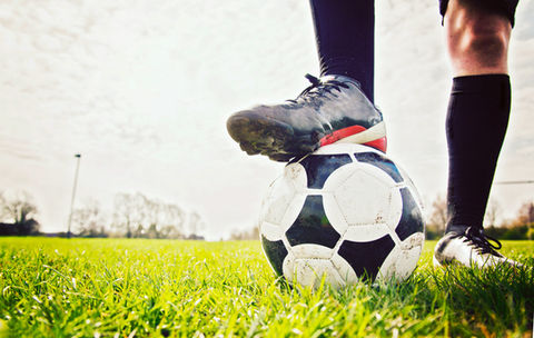 The image shows a football player with one foot resting on a classic black-and-white soccer ball on a green grass field.