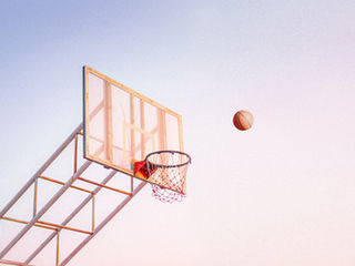 A basketball is soaring towards the basket against a soft blue and pink sky.