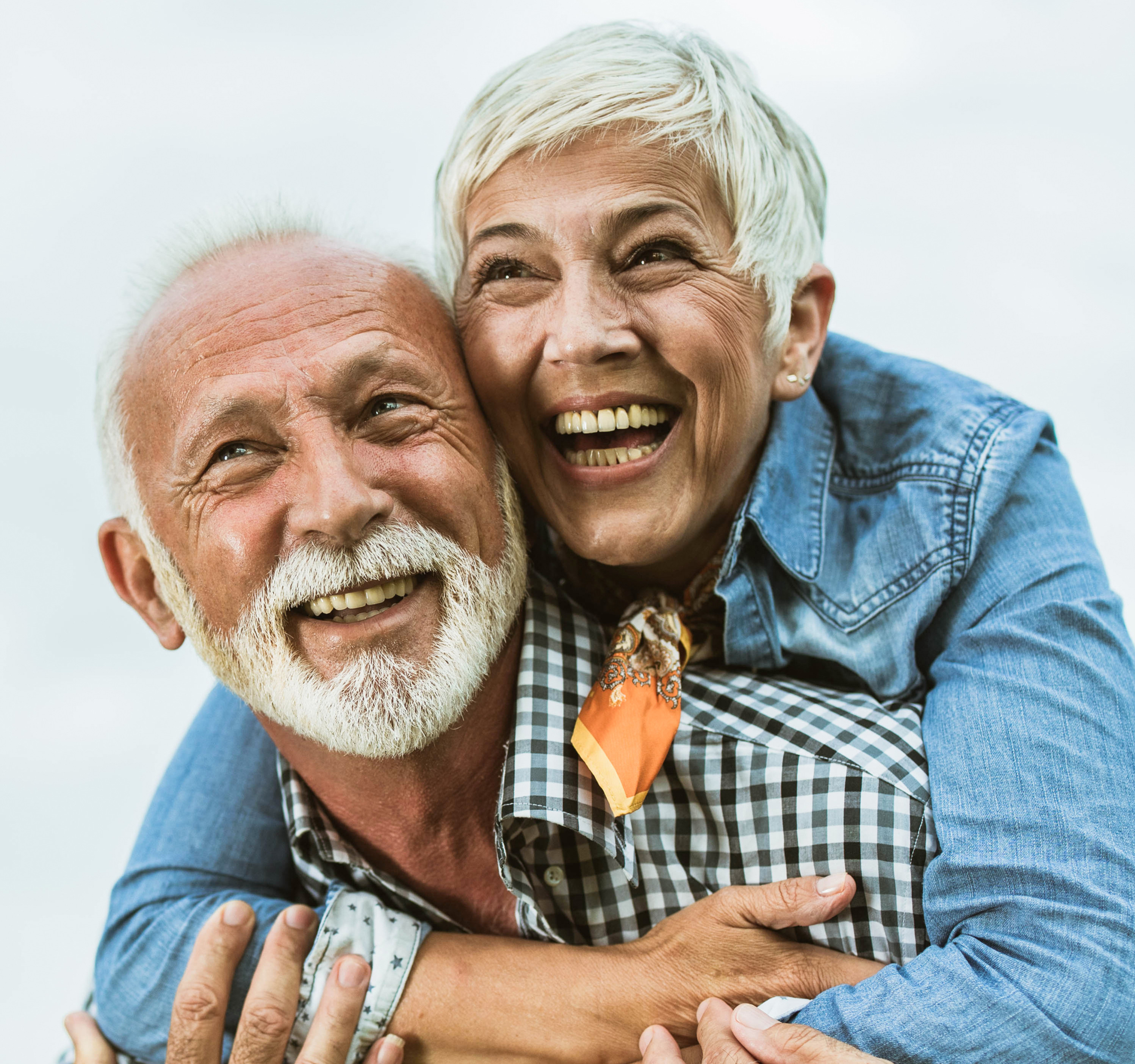 Smiling elderly couple hugging, woman on man’s back; both laughing against a bright white background, wearing plaid and denim.