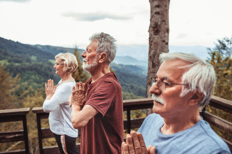 Three older adults practice yoga outdoors, hands in prayer position. They're on a wooden deck with a scenic mountain view, in a serene atmosphere.