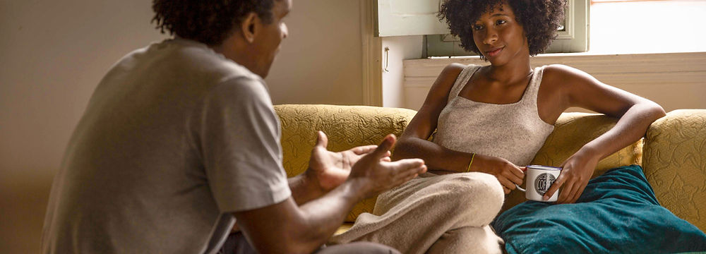 Woman sitting on a couch facing her husband as he tries to explain how he feels during their conversation.