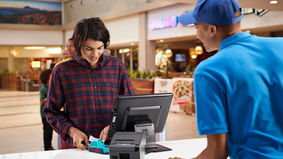 A customer cheerfully completes a transaction at a mall kiosk, interacting with a friendly employee in a blue uniform.