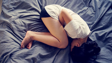 Woman curled up on a blue bed, face covered by a white pillow, suggesting stress or sleep.