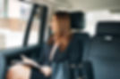 A woman in business attire sits in the back seat of a car, holding a notepad and pen, looking thoughtfully out the window