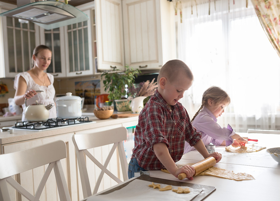 A mother stands at the stove and looks toward her son and daughter as they roll cookie dough together at the kitchen table