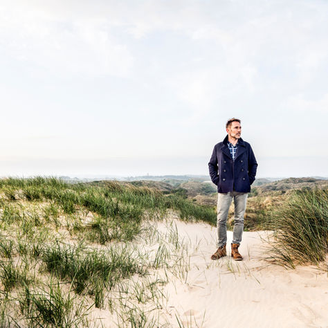 A man in his 50s stands alone on coastal dunes, hands in pockets, looking toward the horizon on an overcast day.