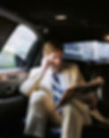 A businessman reading a newspaper in the backseat of a car while using a mobile phone