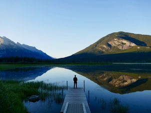 A person looks out into nature and the mountains reflect on the lake.