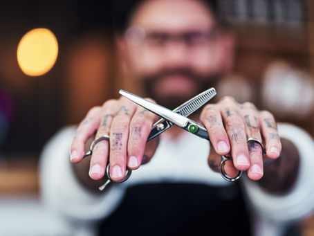 Tattooed hands holding scissors in a blurred barber shop, with warm lights in the background, conveying a focused, professional mood.