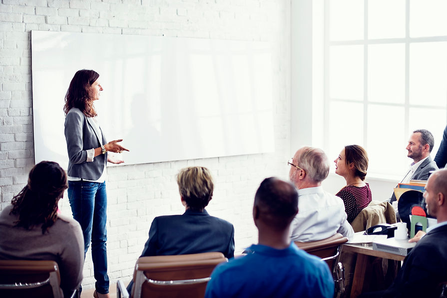 A woman stands in front of a blank whiteboard, gesturing as she presents to a seated group