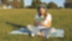 Young woman sitting on grass in park, wearing blue headphones, looking at a tablet. She's on a blanket, wearing a white shirt and jeans.