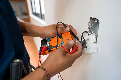 Hands using an orange digital multimeter to test a white wall plug socket