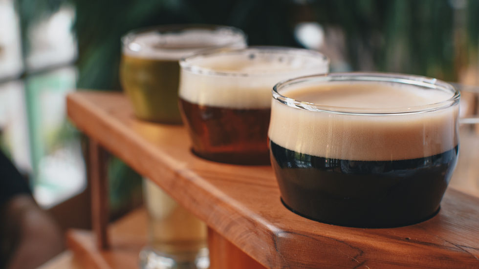 Three glasses of stout beer on a wooden tray, each with different colours and foam. The background is blurred greenery, creating a relaxed mood.