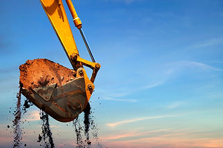 Close-up of a yellow excavator bucket with soil dropping from it against a blue and pink sky