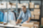 A man standing at a desk smiling, processing shipment orders on his laptop, surrounded by boxes