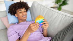 Smiling person in a purple shirt relaxes on a gray sofa, holding a rainbow-colored pop-it toy. Soft light, indoor setting, relaxed mood.