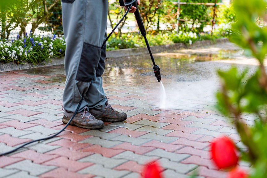Person in gray pants pressure washes colorful brick pathway surrounded by greenery and flowers