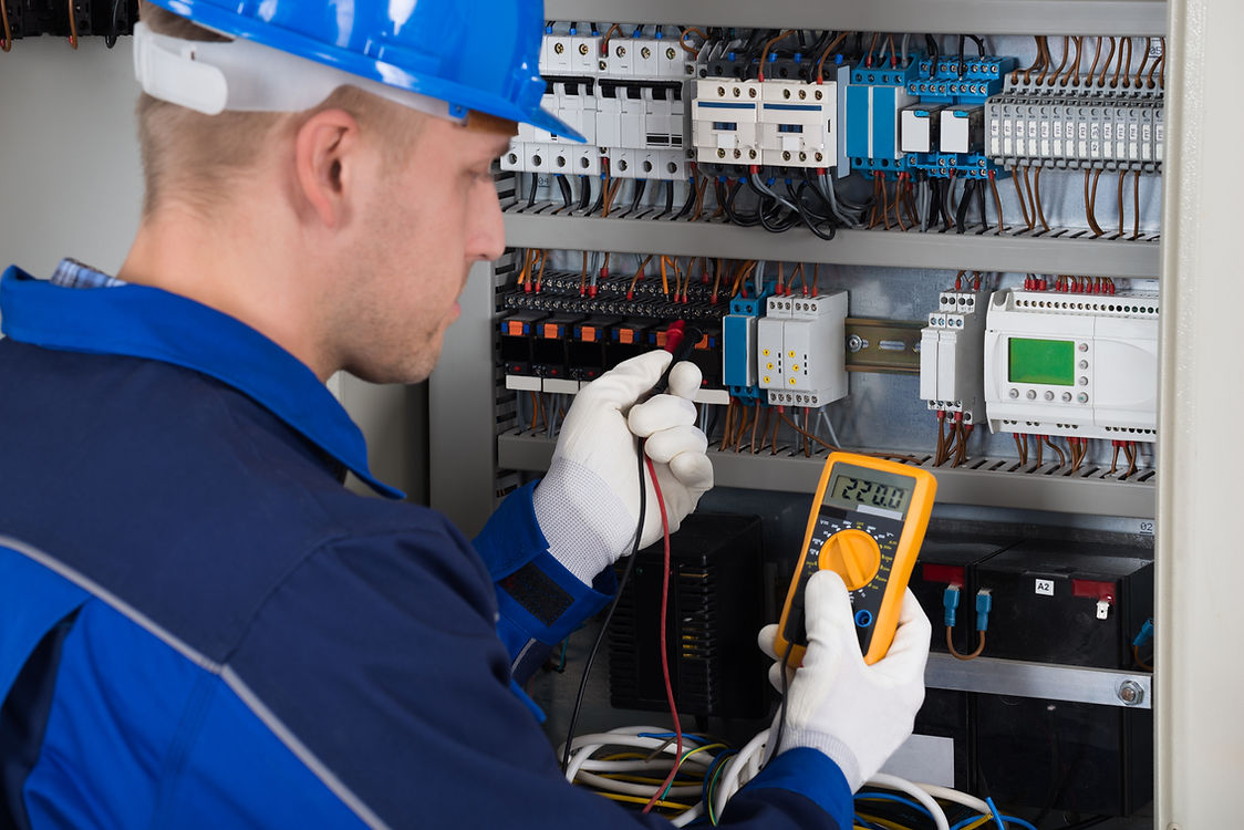 An electrician in blue overalls and gloves checks a circuit panel with a digital multimeter
