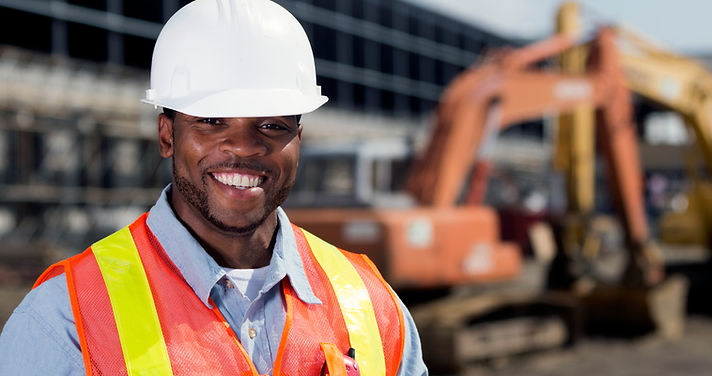 Construction Site Supervisor smiling whilst wearing a white hard hat and orange vest with construction equipment behind him.