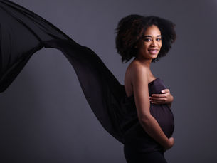Smiling pregnant woman in a dark studio, wrapped in flowing black fabric against a gray background.