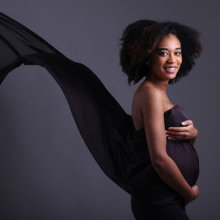 Smiling pregnant woman in a dark studio, wrapped in flowing black fabric against a gray background.