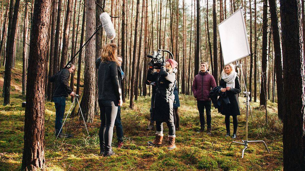 Film crew in a forest setting with trees. A camerawoman films a woman, sound technician holds boom mic, and others watch. Bright, natural lighting.
