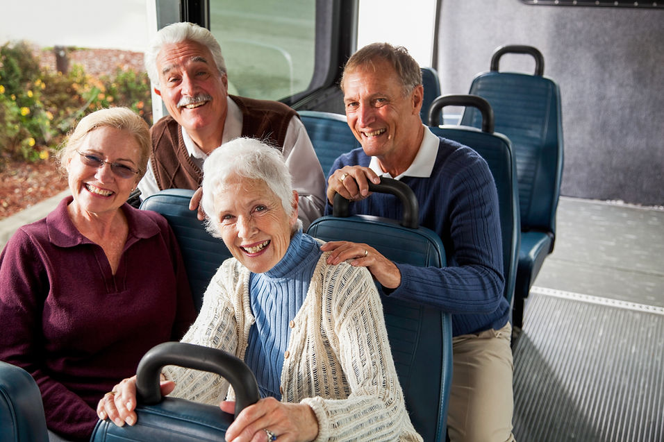 A group of four senior citizens riding in a van or shuttle bus, smiling at the camera.  Th