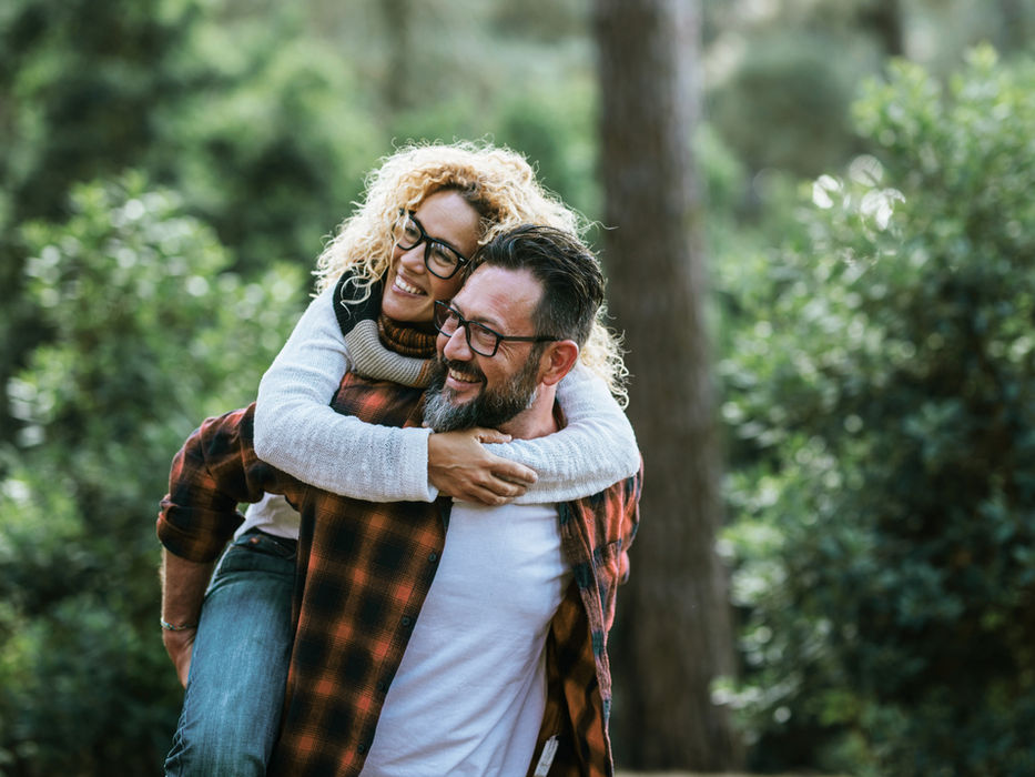 Woman with glasses being carried piggyback by a man in the forest, both smiling.