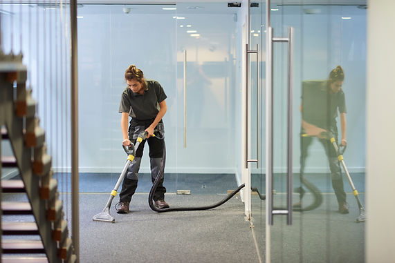 A person in work attire is vacuuming a carpeted hallway, reflected in the glass walls