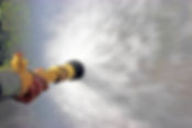 A hand grips a brass fire hose nozzle, spraying a powerful jet of water, creating a dynamic spray pattern