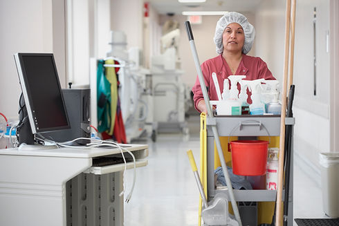 Custodian in red uniform pushes cleaning cart down bright hospital corridor