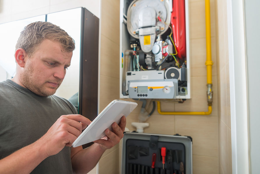 A man checks a tablet in a utility room, focused on a wall-mounted boiler system
