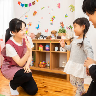 A teacher warmly greets a young girl and her parent in a cheerful classroom decorated with toys and colorful wall art.