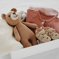 a pile of neatly folded baby clothes, baby shoes, and a brown stuffed bear
