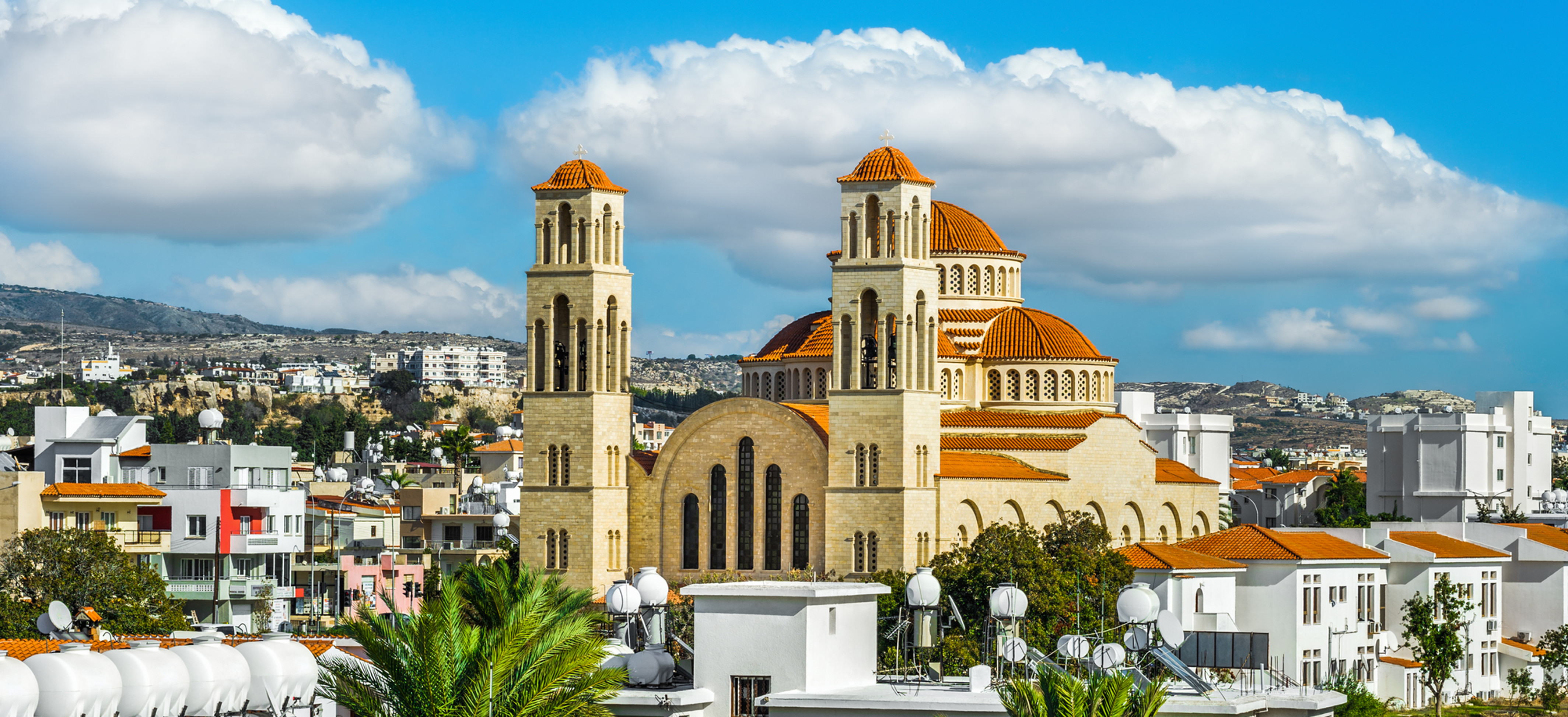 Large stone church with orange domes and twin towers above white buildings and palm trees under bright blue sky and clouds