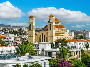 Panoramic view of a large Byzantine-style church with twin bell towers and orange domes, surrounded by modern homes and palm trees under a bright blue sky in Cyprus.